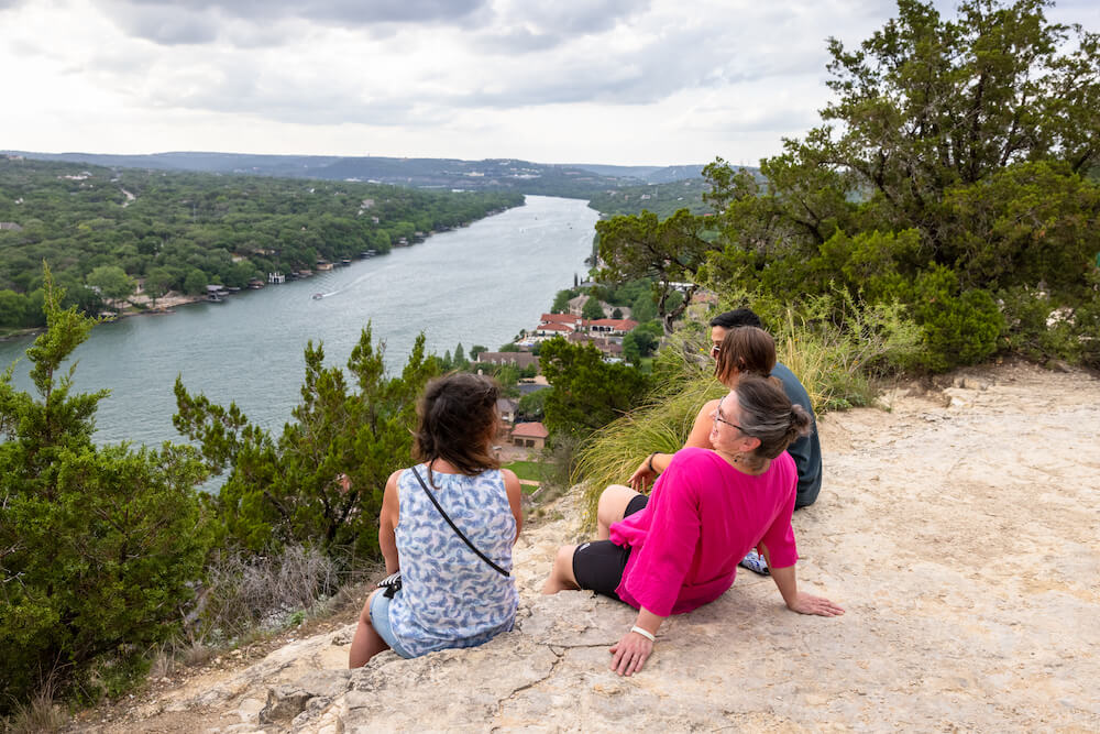 Three people sitting at the top of a hill overlooking trees and a river.