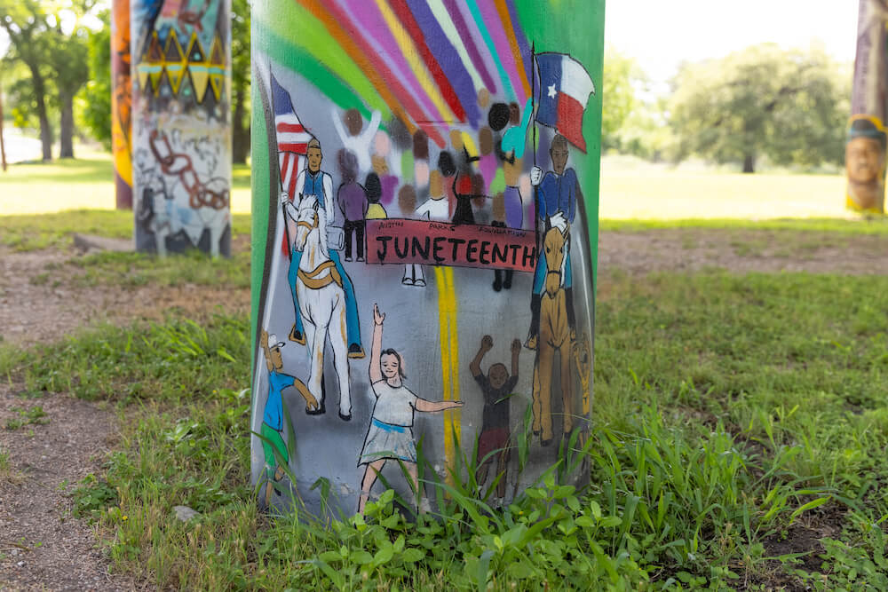 Juneteenth mural on a cement pillar depicting two Black men on horseback, one carrying the Texas flag and the other carrying a US flag. In front of them, there are children in the street, and a large crowd of people behind them with a rainbow overhead.