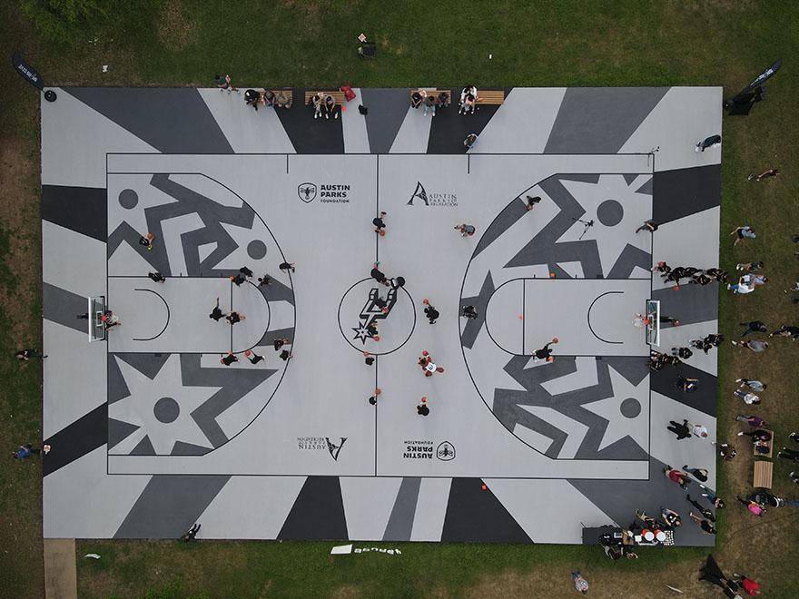 An overhead shot of a basketball court painted with a gray and black geometric pattern around the edges. The middle displays a San Antonio Spurs logo with Austin Parks Foundation and Austin Parks and Recreation Department Logos on either sides. Children play with basketballs across the court.